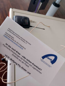 Ein Schreiben, auf dem der Titel der Veranstaltung steht, liegt auf einem Tisch. Der Titel lautet: Ist das noch Krise - oder schon Krankheit?