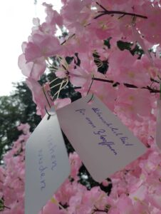 zwei Zettel sind an einen Baum gehängt, die künstliche Kirschblüten trägt (Wunschbaum). Auf den Zetteln stehen Wünsche.
